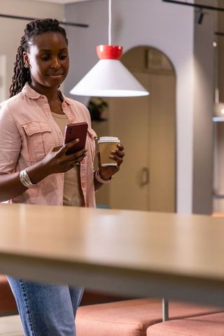 African american woman checking smartphone while holding coffee in modern coworking lounge