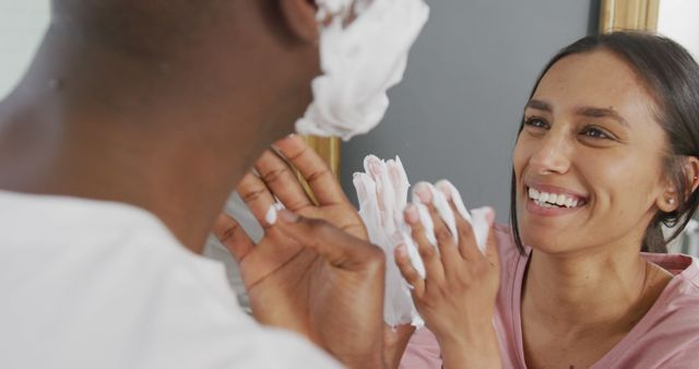 Young couple having playful moments while getting ready in the bathroom. The woman is smiling and rubbing shaving cream on her partner's face. Captures a mix of fun, love, and intimacy. Ideal for advertising personal care products, morning routines, and healthy relationship dynamics. Perfect visual for lifestyle blogs and romantic comedies.