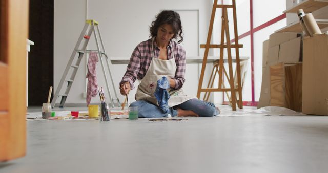 Female artist sitting on floor painting in art studio, surrounded by art supplies and canvases. Can be used for topics on creativity, inspiration, art workshops, or home studio setups.