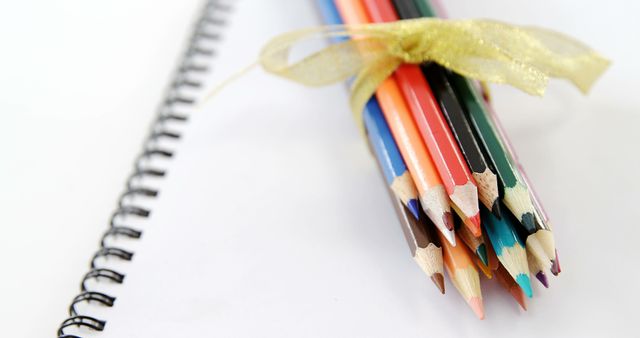 Color pencils tied with a decorative ribbon laying on a clean, white spiral notebook page. Suitable for education-themed visuals, back-to-school advertisements, art workshops, and creativity-focused content.
