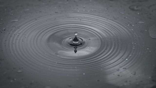 Water droplet forming rising column and creating concentric ripples on calm gray surface