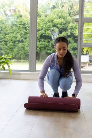 Woman rolling yoga mat at home near sunlit window for at-home workout and mindful warmup