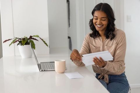 Smiling woman sorting mail at bright kitchen counter with laptop, mug and indoor plant