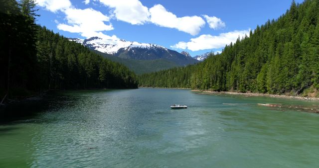 Picture showcases a peaceful mountain lake surrounded by dense evergreen forest with a small boat floating in the serene waters. Snow-capped mountains are visible in the backdrop. This is perfect for travel brochures, nature blogs, and outdoor adventure promotions highlighting remote, beautiful landscapes.
