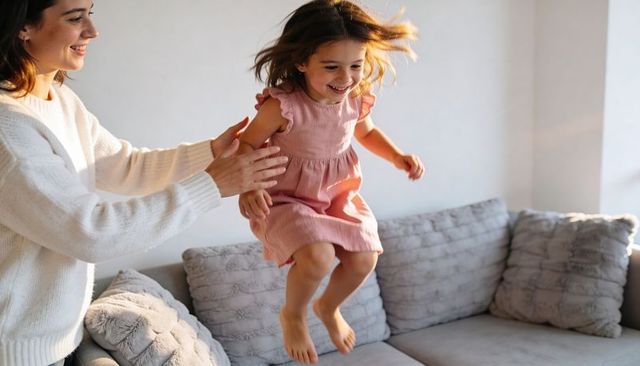 Mother catching laughing little girl jumping on sofa in sunlit cozy living room
