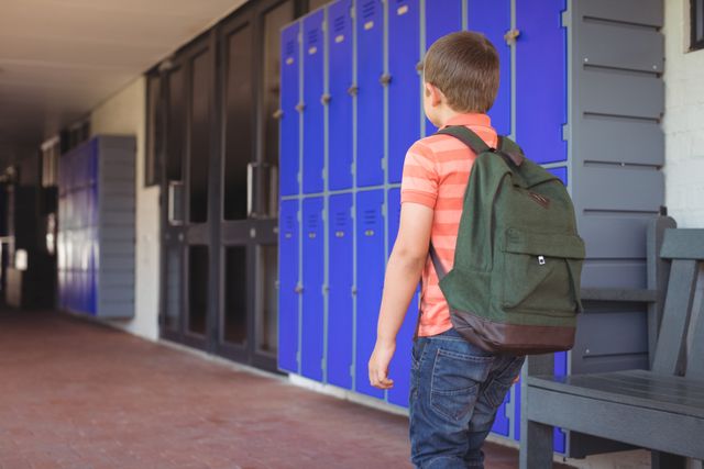 Young boy in casual clothes with backpack walking in school corridor near blue lockers. Ideal for educational materials, back to school promotions, youth and childhood-related content, and school-themed advertising.