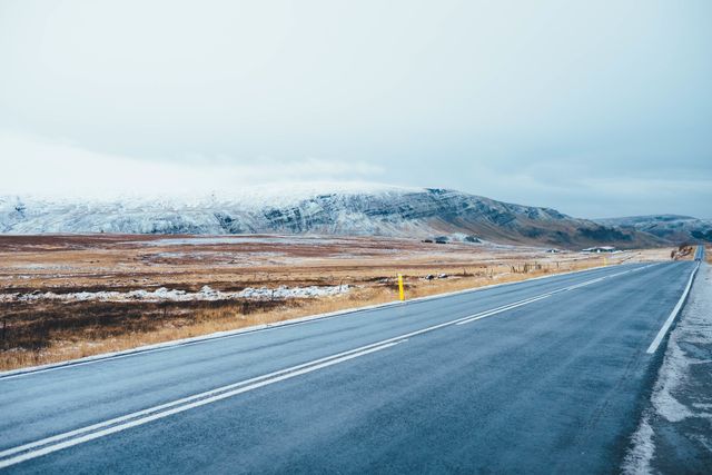 A quiet highway stretches into the distance through a snow-dusted landscape, creating a sense of solitude and adventure. The icy road and the bare, open countryside emphasize the remote and untouched nature. Ideal for depicting winter travel, serene getaway destinations, or themes of isolation and exploration.