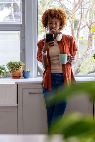 African american woman holding mug and smartphone in kitchen with headphones
