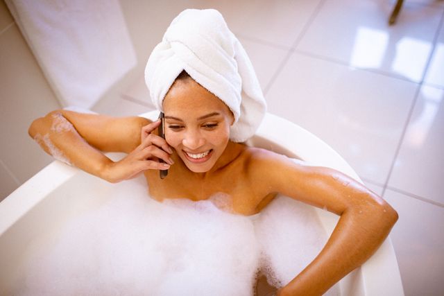 Biracial woman enjoying a relaxing bath with foam while talking on her smartphone. Ideal for concepts related to self care, relaxation, domestic lifestyle, and leisure time at home. Perfect for wellness and lifestyle blogs, advertisements for bath products, and articles on personal well-being.