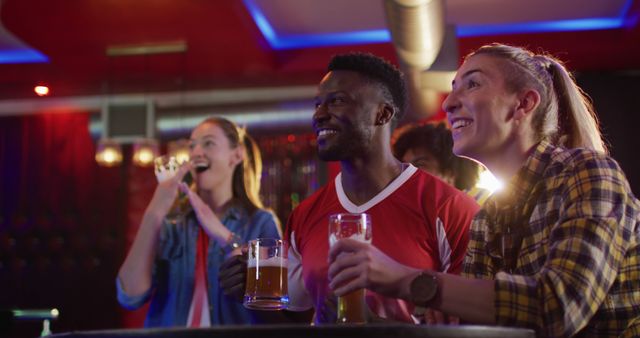 Group of friends holding beer mugs and watching TV in a lively sports bar. Engaging in a fun night out. Suitable for content related to friendship, leisure activities, nightlife scenes, beer advertisements, and socializing in a group setting.