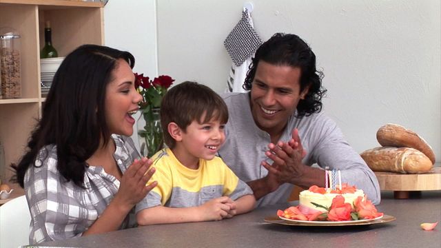 Family gathered in kitchen joyfully celebrating child's birthday. Parents clapping and smiling while child sits in front of birthday cake decorated with flowers. Perfect for ads or articles about family bonding, birthday celebrations at home, or parenting tips.