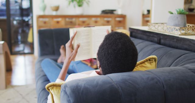 Happy african american woman lying on sofa in living room, reading book. domestic life, spending time at home.