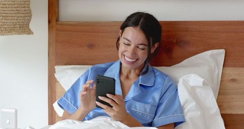 Woman in blue satin pajamas enjoying smartphone leisure on bed