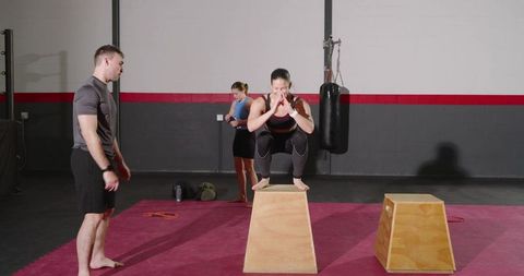 Female athlete performing box squat on plyo box with trainer spotting during group training session