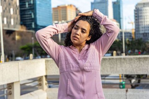 Young woman adjusting curly hair on urban overpass wearing pink zip-up jacket and rings
