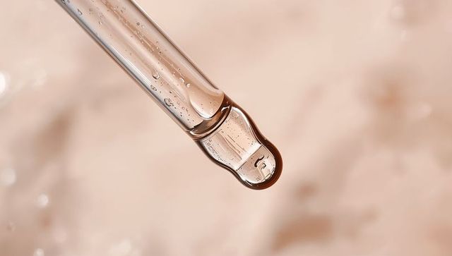Glass dropper releasing clear serum droplet with micro bubbles on soft neutral background