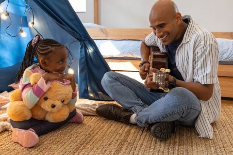 Diverse dad and daughter playing guitar in cozy bedroom tent hugging teddy bear smiling