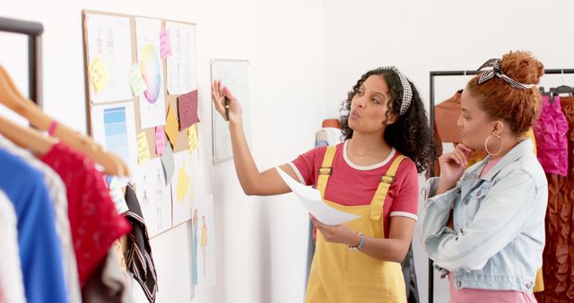 Image of two young fashion designers brainstorming new collection concepts in a modern studio. They are discussing ideas pinned to a concept board. This image can be used for articles or marketing materials related to fashion, creative collaboration, design processes, and teamwork in the fashion industry.