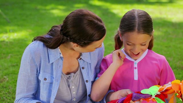 A delighted young girl opening a colorful kite while sitting beside her mother in a park. This scene conveys family bonding and joy in a sunny outdoor environment. Ideal for illustrating family moments, parental love, and the beauty of parent-child interactions. Suitable for parenting resources, recreational activity guides, or family lifestyle branding.