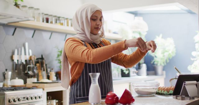 Image of happy biracial woman in hijab baking in kitchen sprinkling flour and using tablet. Happiness, communication, health, inclusivity and domestic life.