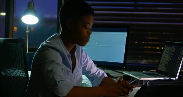 African American woman focusing on computer screen late at night in office. Monitors displaying code and task workload, indicating she is engaged in programming or IT-related tasks. Setting includes desk lamp providing illumination. Useful for depicting night shifts, dedication in tech industry, hardworking professionals, or work-life balance scenarios.