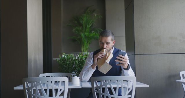 Caucasian businessman using smartphone and eating croissant at table in cafe, copy space. Lifestyle, communication, cafe, business and city, unaltered.