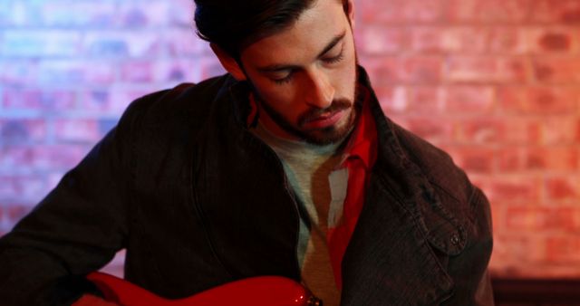 Young man playing an electric guitar against a brick wall background. He seems to be focused on his music and is wearing casual clothing. Ideal for music-related advertisements, websites promoting musicians and artists, or any context highlighting music passion and talent.