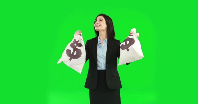 A young Caucasian businesswoman looks up with a smile, holding bags with dollar signs on them, against a green screen background, with copy space. Her expression and the money bags suggest a theme of financial success or wealth.