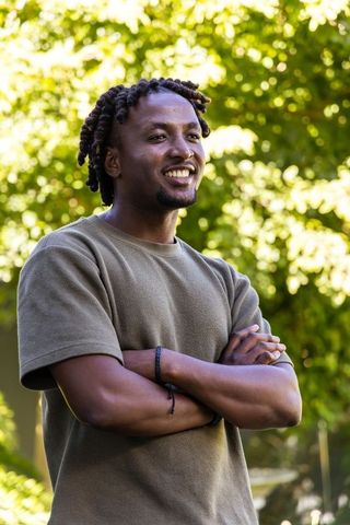 African american man standing in sunlit park smiling with arms crossed wearing olive shirt