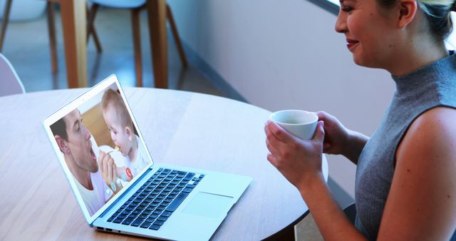 Woman sitting at table, video calling family on laptop, holding coffee cup, smiling happily. Perfect for themes on communication, remote interactions, family bonding, and modern technology. Useful for articles, blogs, and advertisements depicting remote connections and staying in touch with loved ones.