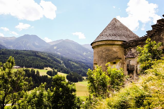 This depicts an ancient stone tower overlooking a beautiful lush mountain valley on a bright summer day. The old tower juxtaposes the serene natural landscape, creating a perfect balance between history and nature. Ideal for use in travel and tourism promotions, historical education materials, and outdoor adventure advertising.