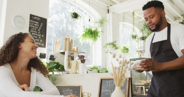 Barista serves coffee to smiling customer in light-filled, cozy cafe adorned with plants. Perfect for use in marketing materials for cafes, hospitality, lifestyle promotions, or social media content focusing on customer service and friendly experiences.