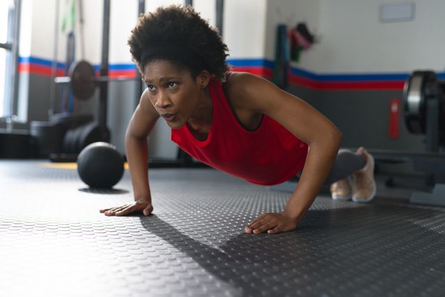 This image depicts an African American woman performing push-ups in a gym. She is wearing a red tank top and has afro hair. The setting includes gym equipment and a rubberized floor. This image can be used for promoting fitness, health, and active lifestyle content. It is ideal for use in advertisements, fitness blogs, workout guides, and health-related articles.