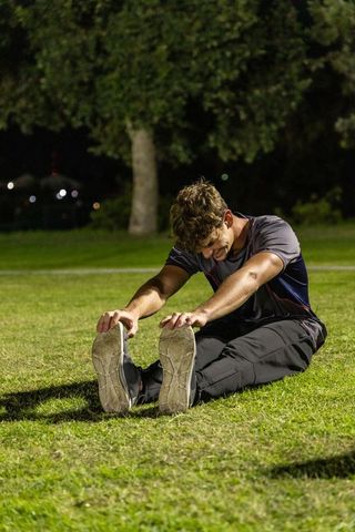 Nighttime park stretching male athlete seated forward bend hamstring stretch on grass