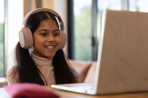 Indian preteen girl wearing headphones smiling at laptop during cozy home learning