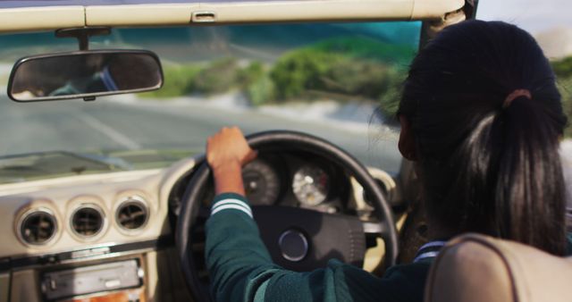 African american woman adjusting rear view mirror while driving along country road in convertible car. road trip travel and adventure concept