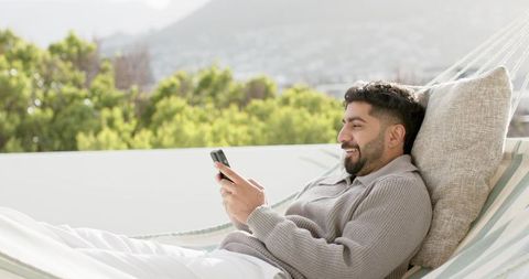 Indian man reclining on striped hammock on rooftop terrace using smartphone, sunny leisure