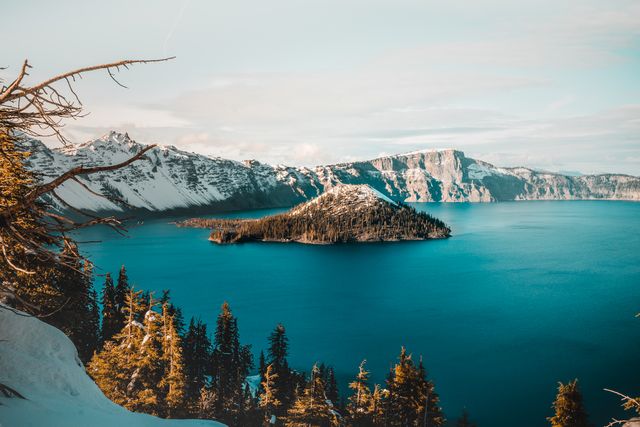 The scenic shot captures Crater Lake with snow-capped mountains and deep blue water, providing a stunning view. Ideal for travel brochures, nature magazines, or outdoor adventure blogs showcasing the beauty of winter landscapes and national parks.