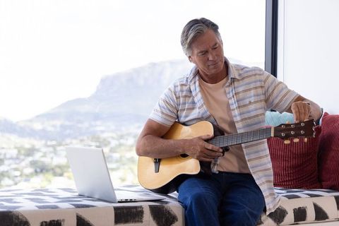 Mature man tuning acoustic guitar near window with laptop and mountain view