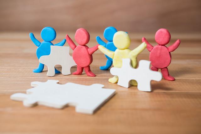 Colorful clay figures standing on a wooden floor, holding jigsaw puzzle pieces, symbolizing teamwork and collaboration. Ideal for illustrating concepts of problem solving, creativity, and group activities in educational or playful contexts. Suitable for use in educational materials, team-building presentations, and creative workshops.