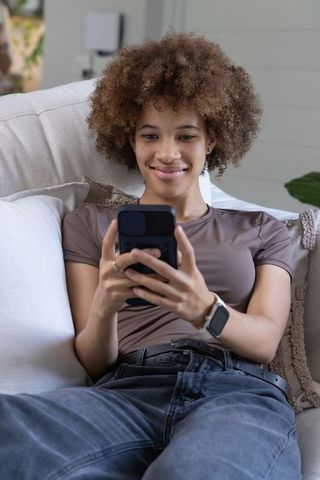 African american woman relaxing on sofa using smartphone and smartwatch in cozy living room