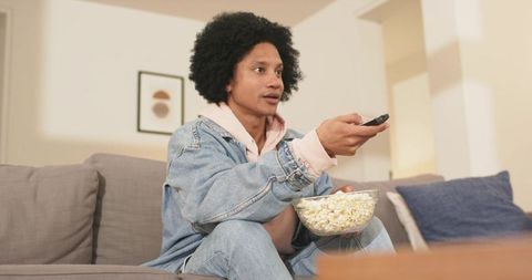 Mid adult african american man relaxing on sofa holding popcorn and remote watching tv