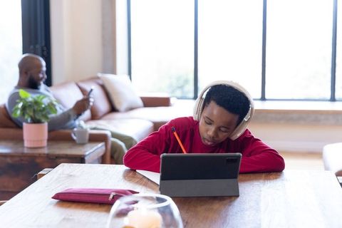 African american boy doing remote learning on tablet with headphones, dad relaxing nearby