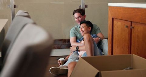 Diverse gay couple relaxing among moving boxes at new home