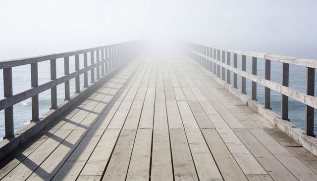 Wooden pier stretching into dense fog over calm sea, minimalist coastal perspective