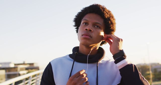 This image shows a young man with an afro hairstyle adjusting his earphones while standing outdoors during sunset. He is wearing a hoodie, and a smartwatch is visible on his wrist. The urban background and the soft dusk light create a relaxed and focused atmosphere. This photo can be used for themes involving youth culture, music, technology, urban lifestyle, and outdoor activities.