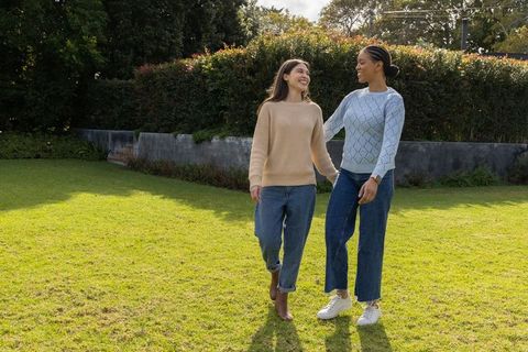 Diverse female friends walking on sunlit lawn wearing sweaters and jeans