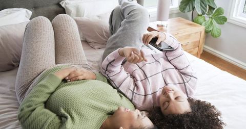 Female friends sharing smartphone and laughing while lying head-to-head on cozy sunlit bed