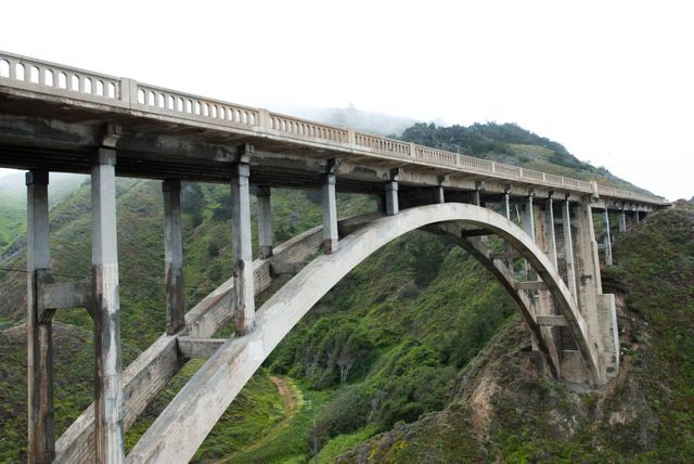 Concrete arch bridge crossing lush green valley, representing feat of historic civil engineering. Ideal for illustrating topics on engineering, transportation, architecture, travel, and scenic routes. Defines structure blending with natural landscape.