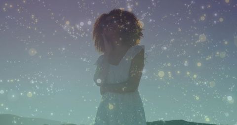 Woman standing at twilight wearing white dress with sparkling bokeh lights and silhouette